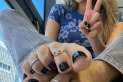 A person sitting cross-legged indoors, showing their black-painted toes adorned with several rings, while giving a peace sign with their hand. They are wearing a blue floral shirt.
