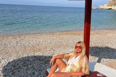 A person relaxing on a beach lounger under a straw umbrella, with a calm sea in the background and rocky cliffs on the side.