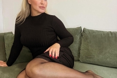 A woman in a black ribbed dress sitting on a green sofa, with her legs crossed and wearing sheer black tights, posed with one hand on her knee and a slight smile.