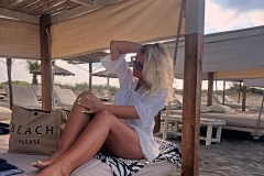 A woman in a white cover-up is relaxing on a beach bed, sitting with her legs crossed. She has sunglasses on and is playing with her hair. A beach bag that reads 'BEACH PLEASE' is next to her.